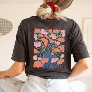 May include: Dark gray t-shirt featuring a colorful floral design on the back. The graphic includes a blue vase with pink and orange flowers. The shirt is a casual, comfortable style.