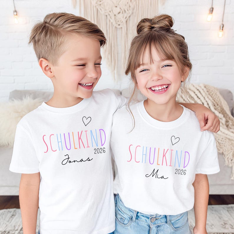 May include: Two white short-sleeved t-shirts featuring the text SCHULKIND in multicolored letters, the year 2026, a small black heart outline, and the names Jonas and Mia printed in black script on the respective shirts.