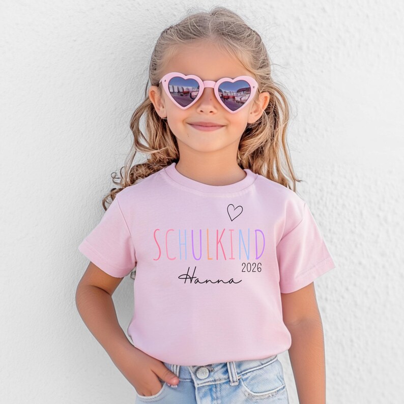 May include: A light pink short-sleeved t-shirt featuring the text SCHULKIND in multicolored letters, a small black heart outline, the name Hanna in black script, and the year 2026. The shirt is paired with light blue denim jeans.