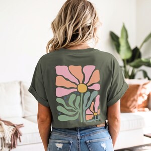 May include: An olive green t-shirt with a floral design on the back. The design features a large flower with pink and orange petals, a yellow centre, and green leaves. The shirt is worn with blue jeans.