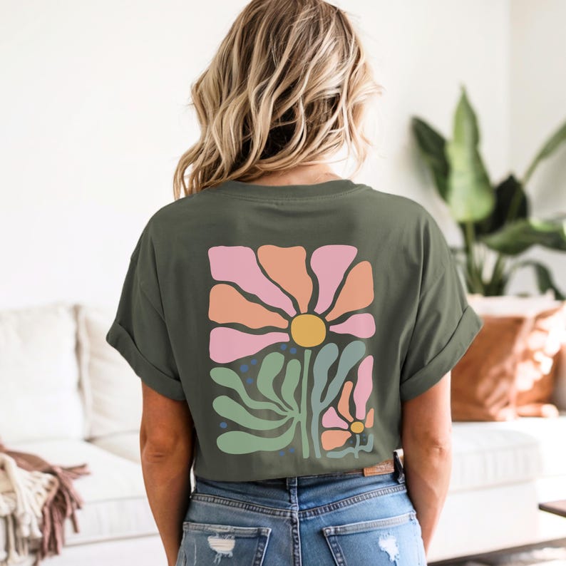 May include: An olive green t-shirt with a floral design on the back. The design features a large flower with pink and orange petals, a yellow centre, and green leaves. The shirt is worn with blue jeans.