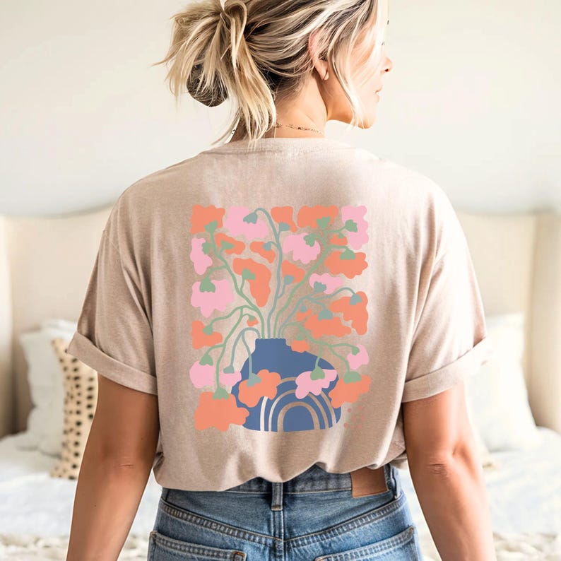 May include: Beige t-shirt with a floral graphic on the back. The design features a blue vase with a rainbow and a bouquet of pink and orange flowers. The shirt is made of a soft material and has rolled-up sleeves.
