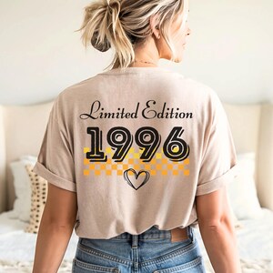 May include: A beige short-sleeve t-shirt featuring the text Limited Edition and the year 1996 in black font. Below the year is a yellow and orange checkered pattern and a black outlined heart graphic centered on the back of the shirt.