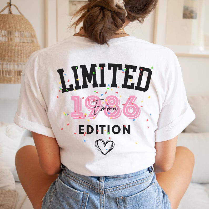 May include: White t-shirt with the text "LIMITED EDITION 1986 Emma" in black and pink, with confetti and a heart graphic. The shirt is a casual style, suitable for everyday wear.