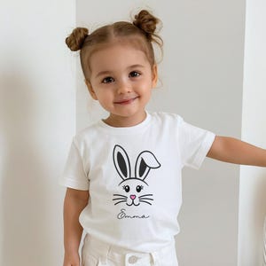 May include: White t-shirt featuring a black and pink bunny face design with the name "Emma" in script. The shirt has short sleeves and is worn by a young child.
