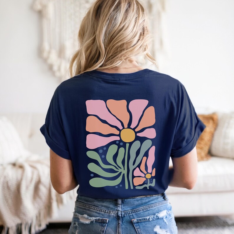 May include: Dark blue t-shirt with a floral design on the back. The design includes a large flower with pink and orange petals, a yellow centre, and green leaves. The shirt is made of soft material.