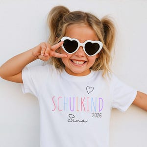 May include: A white short-sleeved t-shirt featuring the word SCHULKIND in multicolored pastel letters, a small black heart outline, the name Sina in black script, and the year 2026 in black text, worn with white heart-shaped sunglasses.