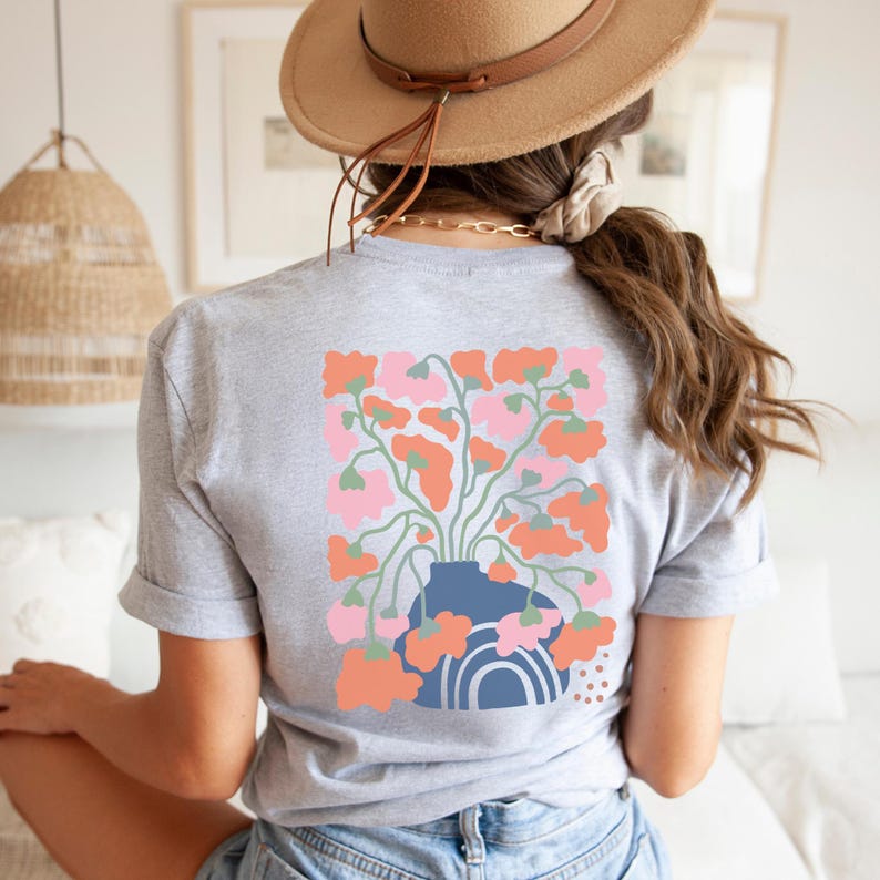 May include: A light gray t-shirt featuring a floral design on the back. The design includes a blue vase with pink and orange flowers. The shirt has short sleeves and a rolled hem. The overall theme is floral and botanical.