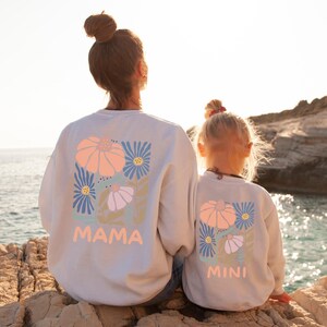May include: Two light gray crewneck sweatshirts featuring a back graphic of stylized flowers in shades of peach, blue, and lavender. The larger sweatshirt has the text MAMA printed in peach, while the smaller sweatshirt has the text MINI printed in peach.