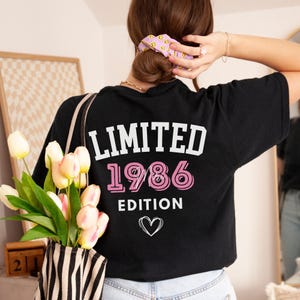 May include: Black t-shirt with the text "LIMITED 1986 EDITION" in white and pink, with a heart graphic. The shirt is being worn by a person with a purple scrunchie in their hair.