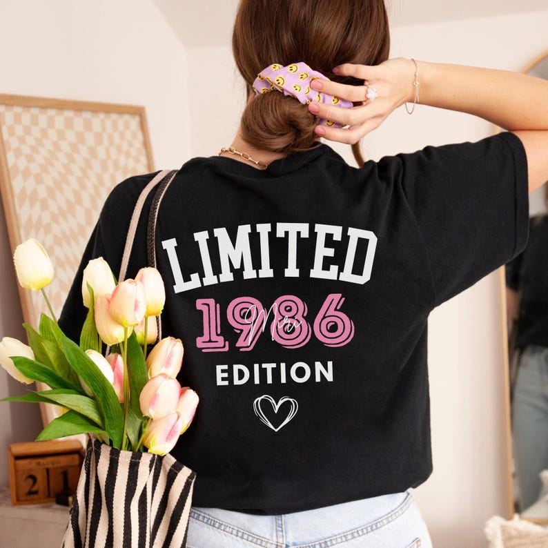 May include: Black t-shirt with the text "LIMITED 1986 EDITION" in white and pink, with a heart graphic. The shirt is being worn by a person with a purple scrunchie in their hair.