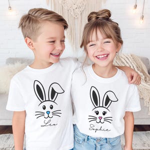 May include: Two children wearing white t-shirts with Easter bunny face designs. The bunny faces are black and white with blue and pink accents. One shirt says "Leo" and the other says "Sophie" in script.