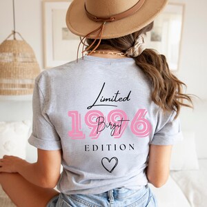 May include: A light grey t-shirt featuring the text "Limited 1996 Birgit Edition" in black and pink, with a black heart graphic. The shirt is being worn.