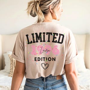 May include: Beige t-shirt with the text "LIMITED EDITION 1986 Laura" in black and pink, with confetti and a heart graphic. The shirt is worn by a person wearing blue jeans.