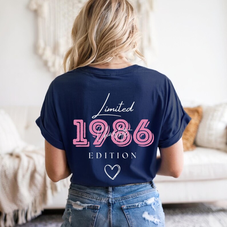 May include: A navy blue t-shirt featuring the text "Limited 1986 Edition" in pink and white script, with a white heart graphic. The shirt has a relaxed fit.