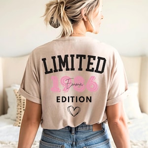 May include: Beige t-shirt with the text "LIMITED EDITION" in black, the number "1986" in pink, and the name "Emma" in cursive pink. A black heart is below the text. The shirt is rolled up at the sleeves.