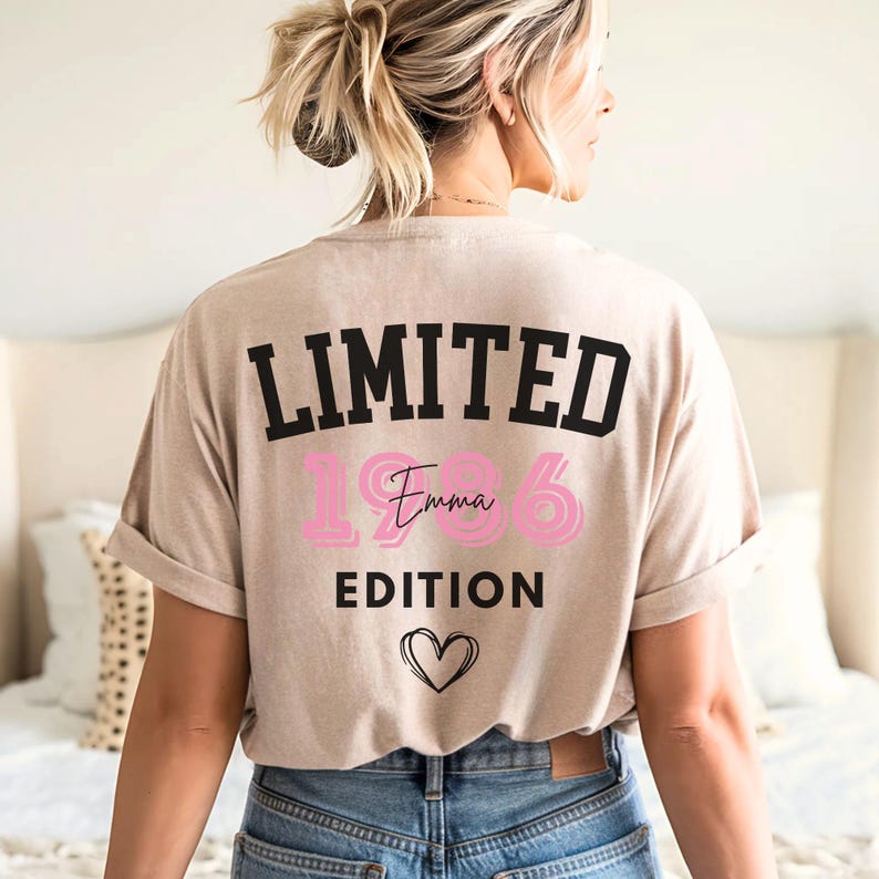 May include: Beige t-shirt with the text "LIMITED EDITION" in black, the number "1986" in pink, and the name "Emma" in cursive pink. A black heart is below the text. The shirt is rolled up at the sleeves.