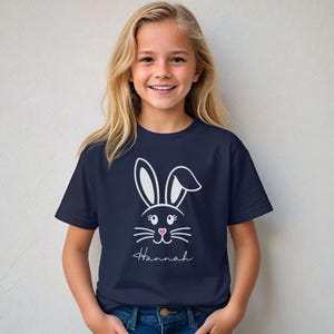 May include: Navy blue t-shirt featuring a white bunny face design with pink nose and whiskers. The name "Hannah" is written in a script font below the bunny face. The shirt is worn by a young girl with blonde hair.