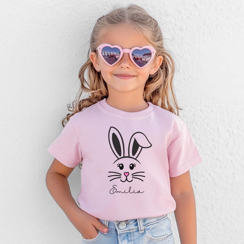 May include: A light pink t-shirt featuring a black and white bunny face design. The bunny has long ears, whiskers, and a pink nose. The name "Emilia" is written in cursive below the bunny face.