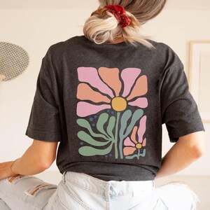 May include: Dark grey t-shirt with a large floral design on the back. The design features a large flower with pink, orange, and yellow petals, green leaves, and smaller flowers. The shirt is made of a soft material.