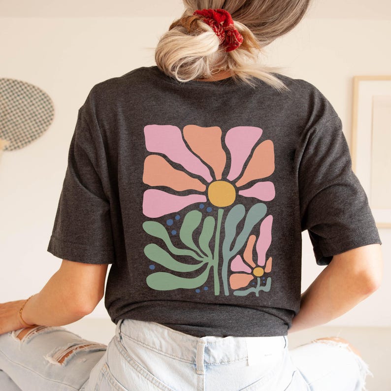 May include: Dark grey t-shirt with a large floral design on the back. The design features a large flower with pink, orange, and yellow petals, green leaves, and smaller flowers. The shirt is made of a soft material.