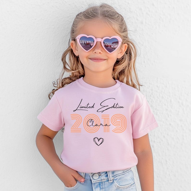 Pu&ograve; includere: Una t-shirt a maniche corte rosa chiaro con la scritta Limited Edition in corsivo nero, l'anno 2019 in numeri a blocchi a righe arancioni, il nome Clara in corsivo nero e un piccolo cuore disegnato a mano in nero sotto il testo.