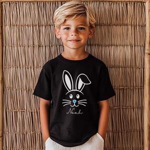 May include: Black t-shirt featuring a white bunny face design with blue accents and the name "Noah" in a script font. The shirt is short-sleeved and suitable for children. The background is a woven bamboo wall.