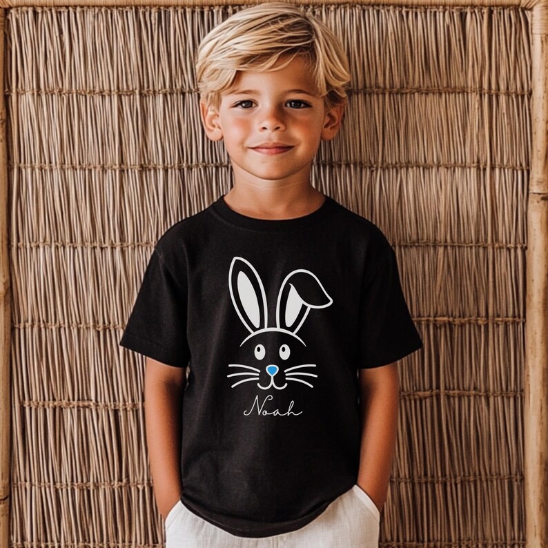 May include: Black t-shirt featuring a white bunny face design with blue accents and the name "Noah" in a script font. The shirt is short-sleeved and suitable for children. The background is a woven bamboo wall.
