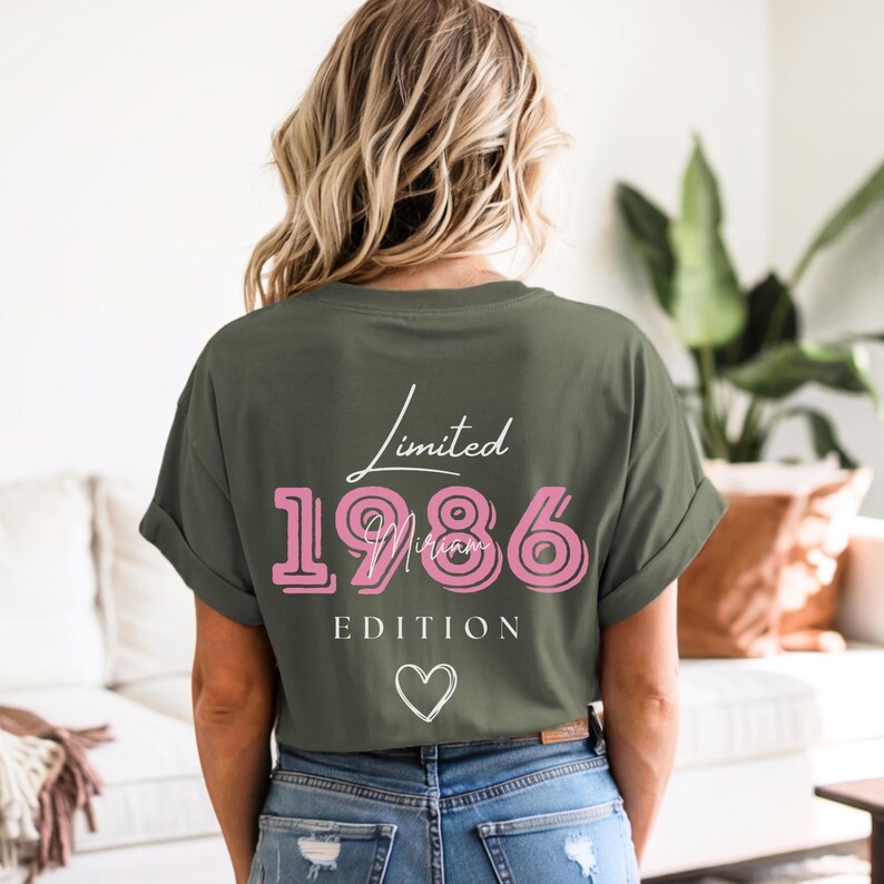 May include: An olive green t-shirt with rolled sleeves. The back of the shirt displays the text "Limited 1986 Edition" in white and pink, with a heart symbol underneath. The shirt is paired with blue jeans.