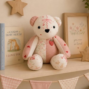 May include: A plush teddy bear constructed from various fabric patches in shades of cream and pink. The bear features a strawberry print, small red hearts, and floral patterns. It has black button eyes, a black nose, and stitched details.