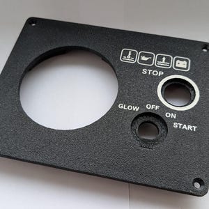 Könnte beinhalten: Schwarzes Bedienfeld mit einer großen kreisförmigen Öffnung und kleineren Löchern für Schalter. Weiße Symbole und Text, darunter "STOP", "GLOW", "OFF", "ON" und "START". Das Panel hat eine strukturierte Oberfläche und Befestigungslöcher.