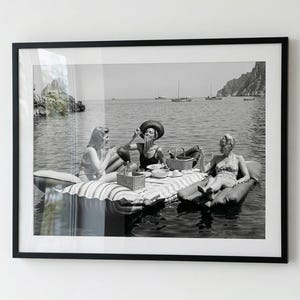 Puede incluir: Impresión enmarcada en blanco y negro de tres personas disfrutando de un picnic en el agua. Están sentadas en balsas inflables, con una cesta de picnic y comida. El fondo muestra barcos y una costa.