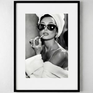 May include: Black and white framed print of a woman applying lipstick. She wears sunglasses, hoop earrings, and a white towel on her head. The woman is also wearing a white robe.