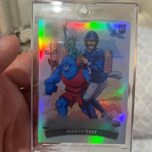 May include: A Jaxon Dart rookie football card in a protective case. The card features a cartoon illustration of a football player and a blue monster with the Statue of Liberty in the background. The card has a holographic finish.