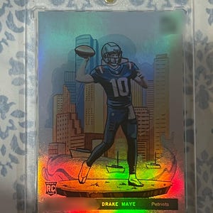 May include: A holographic trading card featuring a stylized illustration of a football player in a Patriots uniform throwing a football. The card is encased in a clear protective holder. The card reads "Drake Maye Patriots".