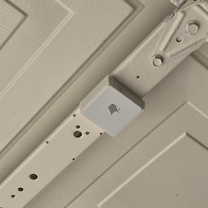 Puede incluir: Un dispositivo rectangular blanco con un logotipo está montado en un riel de puerta de garaje de metal blanco. El riel está sujeto a una puerta de garaje blanca. El dispositivo parece ser un accesorio para el hogar inteligente.