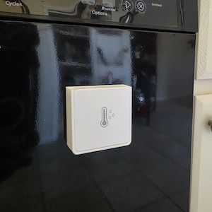 Puede incluir: Un panel de control blanco y cuadrado con un icono de termómetro y gota de agua está adherido a un electrodoméstico negro. El electrodoméstico tiene botones etiquetados como "Heavy Normal", "Soak & Clean", "Heat Dry", "Sani Rinse" y "Start Cancel".