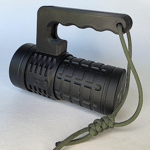 Puede incluir: Una linterna negra con un cuerpo texturizado y un asa incorporada. El asa es de color gris oscuro y tiene un lazo para un cordón verde. El cordón está anudado y cuelga. La linterna tiene un puerto USB.