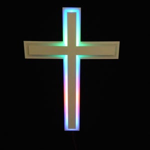 May include: A white cross with a colorful LED light border. The cross has a rectangular design with a multi-colored light border of blue, green, and red. The background is black.