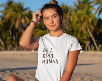 May include: A white t-shirt with the text "BE A KIND HUMAN" in black. The person is sitting on a beach with palm trees in the background. The person is wearing black shorts and bracelets.