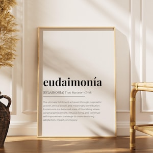 May include: A framed print with the word "eudaimonía" in a bold, black font. Below, the definition is provided in smaller text. The print is in a gold frame and leans against a white wall. A decorative vase and dried plants are in the foreground.