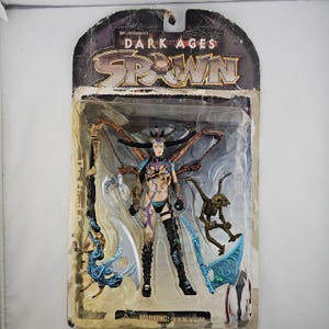 Op de afbeelding: Een Spawn Dark Ages actiefiguur in de originele verpakking. De figuur is van een vrouwelijk personage met een gedetailleerd kostuum, vleugels en accessoires. De verpakking heeft het Spawn-logo en de woorden "Dark Ages".