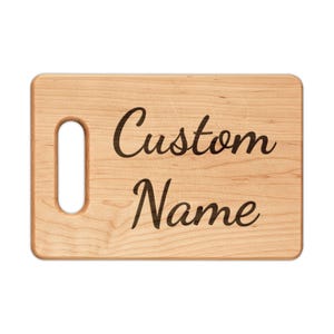 May include: A light-colored wooden cutting board with a handle on the left side. The words "Custom Name" are engraved in a cursive font on the board's surface. The board has a rectangular shape with rounded corners.