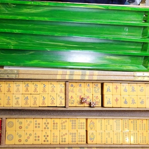 Puede incluir: Un juego de Mahjong presentado en un estuche rectangular con exterior beige texturizado. El set incluye cuatro soportes para fichas de color verde jaspeado y numerosas fichas amarillas con caracteres chinos negros y rojos, símbolos y dos dados marrones.