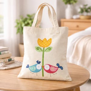 Könnte beinhalten: Cremefarbene Tragetasche mit Blumen- und Vogeldesign. Die Tasche hat eine gelbe Blume mit grünen Blättern und zwei farbenfrohe Vögel. Die Vögel sind blau und rot, mit gemusterten Details. Die Tasche hat zwei Griffe und steht auf einem Holztisch.
