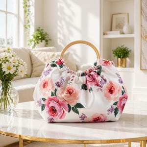 May include: A white handbag with a wooden handle, adorned with a floral pattern of pink and red roses and purple accents. The bag sits on a marble table, showcasing its design and style.