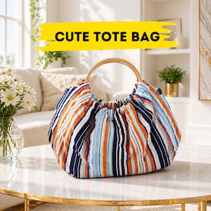 Könnte beinhalten: Eine gestreifte Tragetasche mit Holzgriff. Die Tasche hat vertikale Streifen in Marineblau, Orange und Hellblau. Der Text "CUTE TOTE BAG" wird auf einem gelben Banner über der Tasche angezeigt.