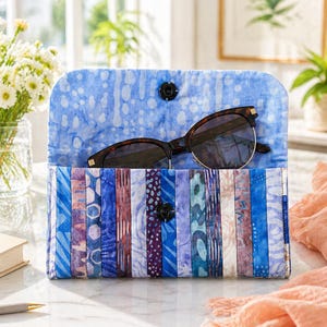 May include: A handmade glasses case with a blue and white patterned interior and a patchwork exterior in shades of blue, purple, and white. The case is open, revealing a pair of tortoiseshell sunglasses. The case is secured with a black snap closure.