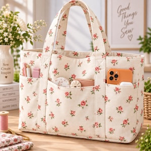 Könnte beinhalten: Eine cremefarbene Canvas-Tragetasche mit einem sich wiederholenden Muster aus kleinen rosa Rosen und grünen Blättern. Die Tasche hat zwei stabile Griffe und drei Außentaschen an der Vorderseite, die ein Smartphone, eine kleine Geldbörse und Nähzubehör enthalten.
