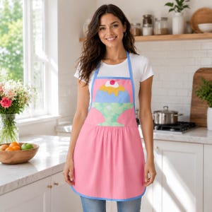 Peut inclure: Un tablier rose avec un motif central représentant une coupe glacée bleue dans un récipient vert surmonté d'une cerise rouge. Le tablier présente un liseré bleu clair sur la sangle du cou et les bords, avec une taille froncée et une jupe rose.